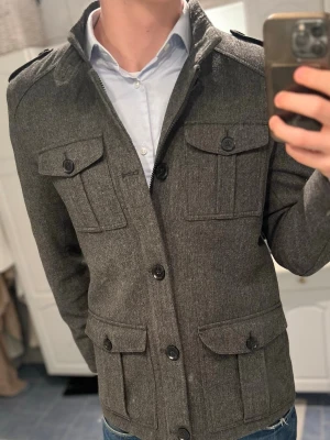 Grå peacoat - Snygg grå jacka med fyra stora fickor framtill och axelklaffar. Stängs med knappar och har en rak passform. Perfekt för dig som gillar en stilren och klassisk look med lite militärkänsla.