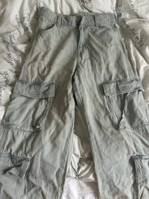 Gråblå cargobyxor med stora fickor - Säljer ett par gråblå cargobyxor med bred passform och flera stora fickor på benen. Byxorna har bälteshällor, knappgylf och bakfickor. Perfekta för en avslappnad och trendig streetwear-look. Materialet känns slitstarkt och passar till många olika stilar.