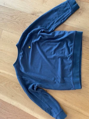 Blå sweatshirt från Lyle & Scott - Grön sweatshirt från Lyle & Scott med klassisk gul logga på bröstet. Tröjan har rund halsringning, ribbade muddar vid ärmslut och nederkant samt en avslappnad passform. Perfekt för en casual look.