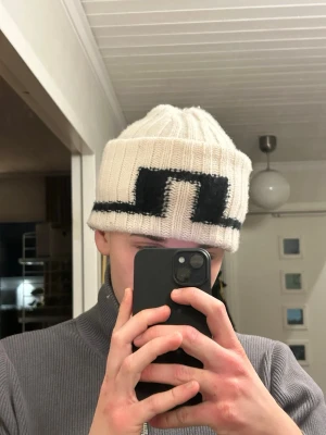 Vit stickad mössa J.Lindeberg - Vit stickad mössa från J.Lindeberg med svart grafiskt mönster framtill. Mössan är ribbstickad och har en uppvikt kant. Tillverkad i mjukt material och passar de flesta tack vare one size-storlek.