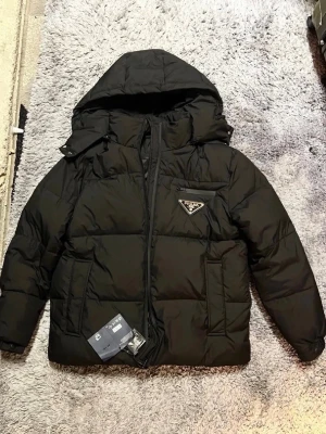 Prada PufferJacka - Säljer en svart pufferjacka från Prada i storlek M. Jackan har huva, dragkedja framtill, en bröstficka med dragkedja och det klassiska Prada-emblemet på bröstet. Fylld med dun för extra varme och har en clean, oversized 