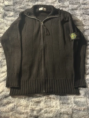 Svart stickad ziptröja från Stone Island - Säljer en svart stickad ziptröja från Stone Island med ribbade muddar och nederkant. Tröjan har hög krage, hel dragkedja framtill och den klassiska Stone Island-loggan på vänster ärm. Materialet är tjockt och strukturerat för extra komfort. Den passar både S och Xs och pris kan självklart diskuteras.