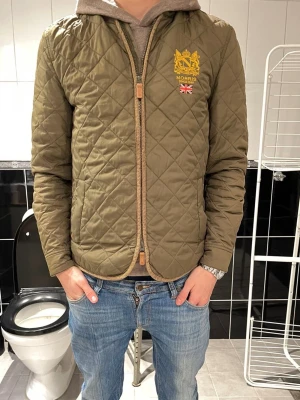 Olivgrön quiltad jacka från Morris - Snygg olivgrön quiltad jacka från Morris med broderad logga och brittisk flagga på bröstet. Jackan har dragkedja framtill och en klassisk, tidlös look. Perfekt för dig som gillar stilrena och bekväma jackor.