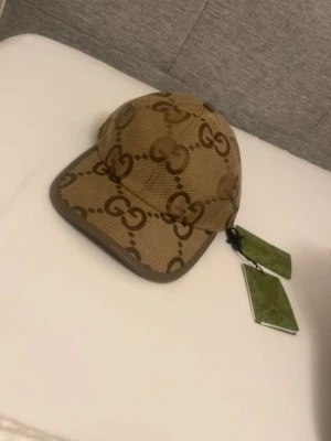 Beige keps med GG-mönster från Gucci - Säljer en beige keps från Gucci med det klassiska GG-monogrammet i brunt över hela kepsen. Kepsen har böjd skärm och bruna detaljer längs kanterna. Tillverkad i canvasmaterial och har originaltaggar kvar. Perfekt för dig som gillar exklusiva accessoarer.