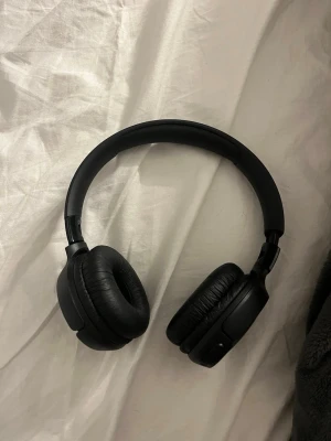 Svarta JBL on-ear hörlurar - Snygga svarta on-ear hörlurar från JBL med vadderade öronkuddar och justerbart huvudband. Trådlös design med knappar på sidan för enkel styrning av musik och samtal. Perfekta för att lyssna på musik eller poddar när du är på språng.