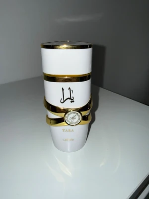 Yara Moi 100ml  - Snygg parfymflaska från Lattafa i modellen Yara. Flaskan är vit med eleganta gulddetaljer och har en unik, staplad design. Dekorerad med arabiska bokstäver och en rund guldetikett. Perfekt för dig som gillar exklusiva dofter och stilren design.