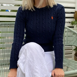 Kabelstickad tröja från Ralph Lauren - Jag säljer den här marinblåa kabelstickade tröjan från Ralph Lauren med ett orange märke. Väldigt bekväm och fin, både att ha den bara som den är men kan också stylas med en skjorta under eller liknande.😊