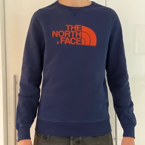 The North Face Sweatshirt Strl: S - En sweatshirt i mörkblå färg med ett orange tryck på bröstet och baksida axel. Den har används en del trots det är den i ett fint skick. Tröjan är lite vintage insperad och därav väldigt fin till alla olika klädstilar!