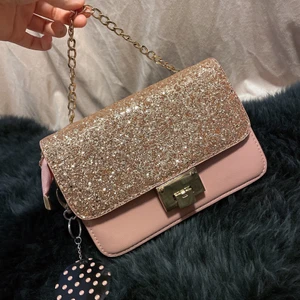 Rosa handväska - En söt rosa väska med glitter och guldiga detaljer. Det kommer också med en spegel som går att sätta på väskan om man skulle vilja det. Den har 3 olika fikor så man får plats med väldig mycket i den dessutom passar den med vilken outfits som helst för att den är så klassiskt. Har bara använt den 1 gång så den är i jätte bra skick!… köparen står för frakten! (Mer bilder kan skickas om önskas)