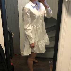 Vit skjortklänning med volanger - Jättefin vit skjortklänning med volanger. Storlek M. Jag är ungefär 173 cm med långa ben och på mig är den lite kortare än knähöjd vilket jag inte känner mig bekväm i. Den är i ett najs krispigt material, känns lyxig!