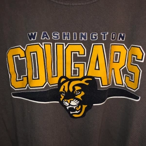 Washington Cougars T-shirt  - Mörkgrå T-Shirt med stort gult tryck på bröstet. Washington Cougars. Uppvikta ärmar. 