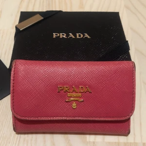 Prada nyckel och korthållare - Nyckel/korthållare från Prada i saffiano läder. Den stängs med tryckknapp och har fack för kort på vardera sida + bakom nycklarna. Finns 5 stycken krokar för nyklar. Som kan ses på bilden har en nyckelkrok lossnat och det finns slitningar på metallen och lädret. Fler bilder kan skickas! Köpt online från Pradas webbsida. Kommer med äkthetsbevis och tillhörande kartong+band+skydd! 