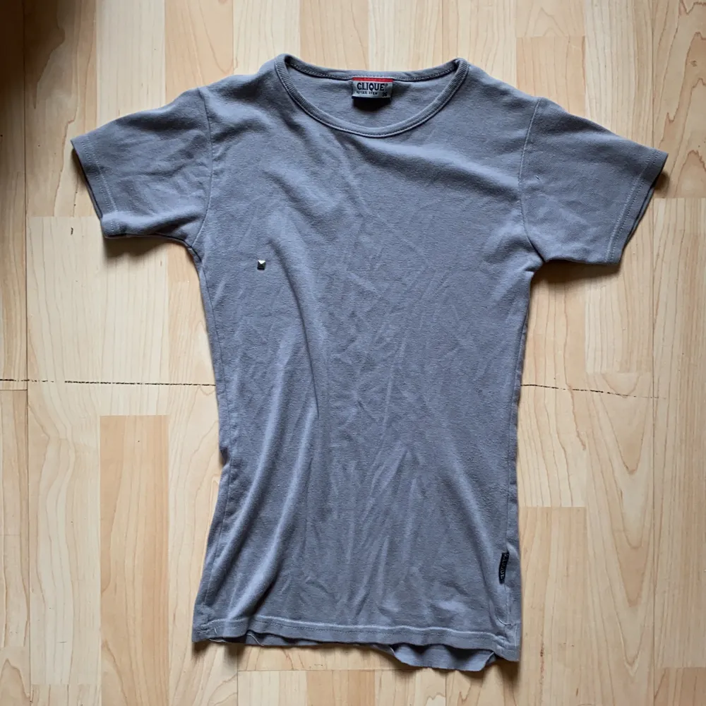 En grå t-shirt i figursmickrande form från CLIQUE. Har endast använts ett fåtal gånger och har en diamant på högra bröstet. Tyvärr, har stygnen gått upp ytterst lite på baksidan men annars är den i utmärkt skick.. T-shirts.