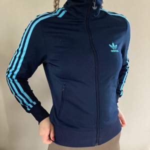 Marinblå ”track jacket” från adidas med turkosa/ljusblå detaljer. Hög krage och fickor. 