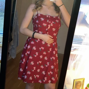Rödblommig klänning - Fin röd sommarklänning med rosa blommor på. Smala axelband med några snygga knappar som detaljer för fram. 