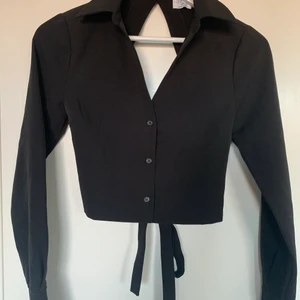 Svart croppad skjorta - Jättesnygg svart croppad skjorta med öppen rygg ifrån na-kd x Hanna Schönbergs kollektion! Den är i storlek 32, jag har vanligtvis 34 och den passar mig men har tyvärr inte kommit till användning❣️ prislappen sitter kvar. Säljer den för 100kr🥰