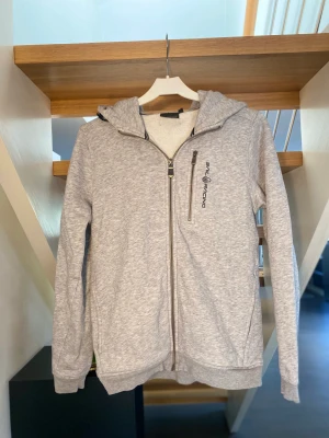 Sail racing jr bowman zip hood - Säljer en ljusgrå Sail racing jr bowman zip hood i storlek 160! Säljes pga för liten! Tröjans luva har dragsko på vardera sida för att kunna anpassa storleken samt en ficka bredvid loggan vid vänster bröst! Nypris 999kr mitt pris 199kr
