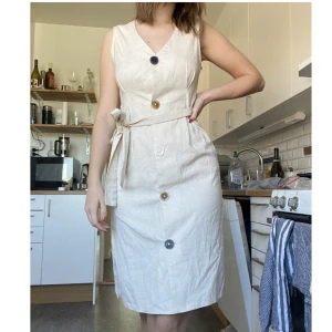 Klänning i linne - Jättefin beige klänning i linne imitation. Köpt utomlands i san fransisco. Olika färger på knapparna.  Aldrig använd  Däremot lite skör i sömmen på ena sidan av klänningen (se bild)