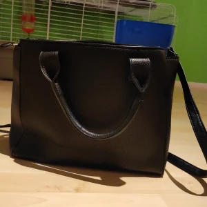 Svart enkel väska - Svart använd men handväska.  På sista bilden kan man se att dragkedjan till sidfickan är något trasig men i övrigt i mycket bra skick.  Den är medelstor men är väldigt rymlig eftersom den har många fickor inuti och utanpå.