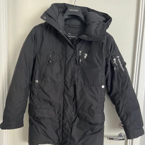 Peak Performance Puffer - Lång dunjacka, lätt figursydd, med flera fickor pä kroppen och ärmarna, och justerbara dragsko. Svart exteriör och helstoppad fuchsia insida. Förstärkningar pà ärmarna. Dragkedja och kardborrestängning. Officiell storlek XS men den passar bättre till en S