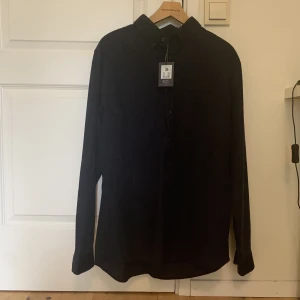 Svart overshirt  - Svart skjorta i manchester-tyg från Dressman. Funkar både som overshirt/överskjorta och som vanlig skjorta. Aldrig använd, prislappen sitter kvar. Storlek XL (43/44). Skicka meddelande vid frågor eller fler bilder 
