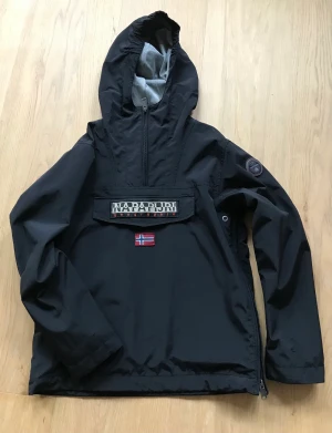Anorak med luva från Napapijri - Svart pull-over anorak med luva från Napapijri i stl 152.   Jackan har en ficka på framsidan och dragkedja upptill samt i sidan. Innerfodret är i mesh och jackan står emot regn och vind.  Nyskick. Endast använd en säsong  