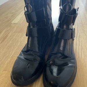 Svarta boots - Säljer ett par coola svarta boots i syntetmaterial med dragkedja och spännen framtill. Perfekta för höst och vinter med en edgy look. De har en robust sula och är i ok  skick. Passar perfekt till både jeans och kjol för en tuff stil.