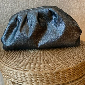 Glitterväska i silver  - En clutch väska från Nelly i silvriga små paljetter. Insidan har ett litet innerfack. Mått bredd 38 cm, höjd 19 cm och ca 11 cm djup. 