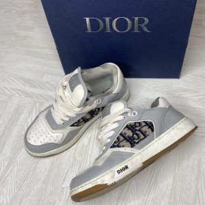Dior B27 Low-Top Sneaker grå & vit - Christian Dior Sneakers 3SN272ZIR vit, grå, beige och blå diorlogga i tyg på sidorna. Stl.36 säljs med låda och dustbag. Nypris 11.500kr kvitto finns från butiken.