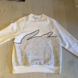 Lacoste sweatshirt  - Vit Lacoste sweatshirt med svarta detaljer. Köpt för någon månad sedan så nyskick 10/10. Även en lite ”osynlig” magficka.