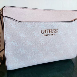 Axelremsväska GUESS - Ganska stor väska från GUESS i ljusrosa. Väskan har ett stort yttre fack och flera mindre fack på insidan.