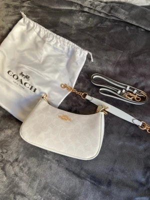 Vit axelremsväska från Coach - Snygg vit handväska från Coach med guldfärgade detaljer och diskret logomönster. Väskan har avtagbar axelrem och medföljer dustbag. Perfekt för dig som gillar stilrena accessoarer med exklusiv känsla.