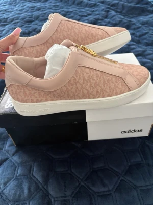 Rosa sneakers från Michael Kors - Snygga rosa sneakers från Michael Kors med guldfärgad dragkedja och diskret logomönster över hela skon. Ovandelen är i textil och syntet med vita sulor. Perfekta för dig som vill ha en clean och trendig look.