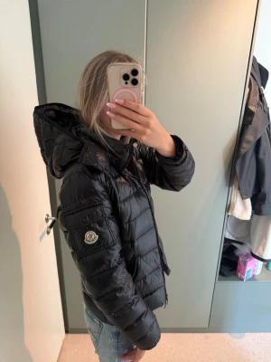 Svart dunjacka från Moncler - Snygg svart dunjacka från Moncler med glansig finish och avtagbar huva. Jackan har Moncler-logga på ärmen, dragkedja framtill och två sidofickor. Perfekt för kalla vinterdagar och riktigt trendig look.