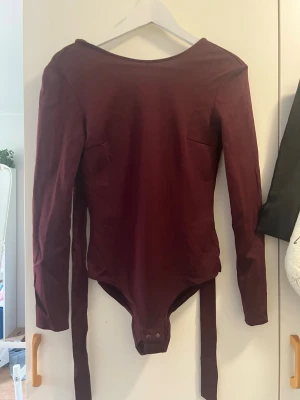 Vinröd långärmad body med band - Säljer en vinröd body med långa ärmar och djup rygg. Bodyn har knäppning i grenen och långa band som kan knytas i midjan eller runt kroppen för en snygg detalj. Materialet ser ut att vara stretchigt och mjukt, perfekt för en trendig look.