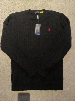 Svart kabelstickad tröja Polo Ralph Lauren - Kabelstickad svart tröja från Polo Ralph Lauren med rund halsringning och den klassiska röda logotypen broderad på bröstet. Tröjan har långa ärmar och ribbade muddar vid ärmslut och nederkant. Perfekt för en stilren och tidlös look.