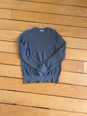 Grå stickad tröja från Zara - Snygg grå stickad tröja från Zara med rund halsringning och ribbade muddar. Tröjan har en enkel, stilren design och är tillverkad i ett mjukt, tunt stickat material som passar perfekt till lager på lager.