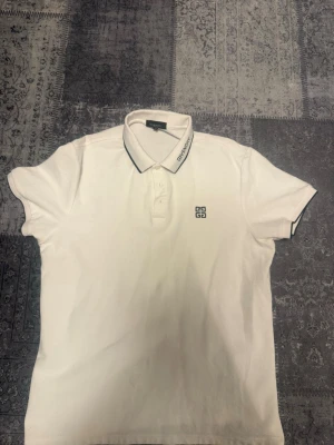 Vit piké med logga och krage - Vit pikétopp med korta ärmar och klassisk krage med diskret gvy logga på bröstet. Kragen har en tunn svart kant och knappslå framtill. Perfekt för en clean och stilren look.