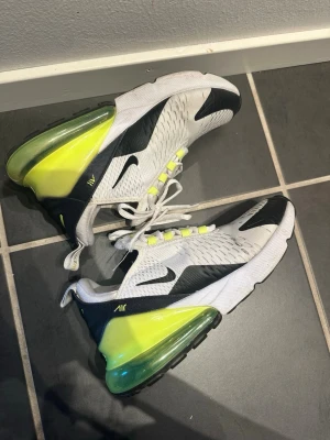 Nike Air Max 270  - Nike Air Max 270 sneakers i vitt meshmaterial med svarta detaljer och neongul/grön häl. Synlig Air-enhet i sulan och klassisk Nike-logga på sidan. Skorna har vita snören och ventilerande ovandel för extra komfort. (Har ett litet hål vid ena tån)
