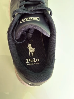 Marinblå sneakers från Polo Ralph Lauren - Stilrena marinblå sneakers från Polo Ralph Lauren med klassisk logga på sidan och plös. Skorna har vita sulor, matchande marinblå skosnören och är tillverkade i slätt lädermaterial. Perfekta för en clean och sportig look.