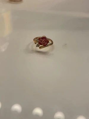 Guldfärgad ring med ros och stenar - Unik guldfärgad ring med en detaljrik ros i rödrosa nyans och små gnistrande stenar på sidan. Ringen har en elegant och romantisk design med blommotiv och glänsande finish. Perfekt för dig som vill ha något extra till din stil. Helt ny