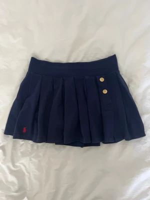 Mörkblå plisserad kjol Polo Ralph Lauren - Mörkblå plisserad kjol från Polo Ralph Lauren med två guldknappar framtill och broderad röd logga. Kjolen har bred midjeresår och är tillverkad i mjukt bomullsmaterial. 