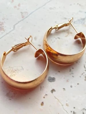 Guldfärgade breda hoops örhängen - Säljer ett par chunky guldfärgade hoops örhängen i metall. De har en bred, rundad form och klassisk klickstängning. Perfekta för dig som gillar statement-smycken och vill addera lite extra bling till din stil. Helt nya och oanvända. Okänt material  ej riktigt guld. 