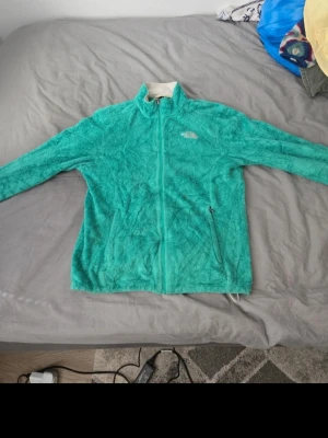 Turkos fleecejacka från The North Face - Mysig turkos fleecejacka från The North Face med dragkedja framtill och två sidofickor med dragkedja. Jackan har hög krage och är tillverkad i mjukt, fluffigt material som håller dig varm. Perfekt för kyliga dagar och enkel att matcha med andra plagg.