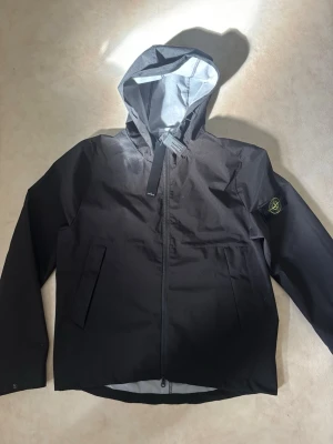 Svart vindjacka med huva Stone island - Snygg svart vindjacka med huva och dragkedja framtill. Jackan har två snedställda fickor och en patch med logotyp på vänster ärm. Insidan av huvan är ljusgrå. Perfekt för blåsiga dagar. Ej använd Storlek L