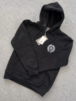 Chrome Hearts Hoodie  - Snygg svart hoodie från Chrome Hearts med stor vit logga och emblem på ryggen samt mindre tryck på bröstet. Klassisk ficka framtill och justerbar huva med snören. Tillverkad i mjukt bomullsmaterial som känns skönt mot huden. Perfekt för en streetwear-look. Har storlek S-XXL