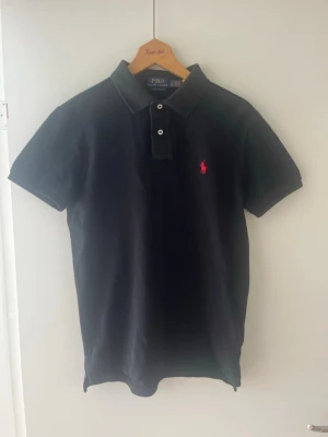 Äkta Svart piké Ralph Lauren - Svart piké från Ralph Lauren med klassisk krage, två knappar och röd broderad logga på bröstet. Storleken är S slim fit och tillverkad i bomull för en snygg look. Köpte den på zalando för 1500 kr och säljer den nu för att den blivit för liten för mig