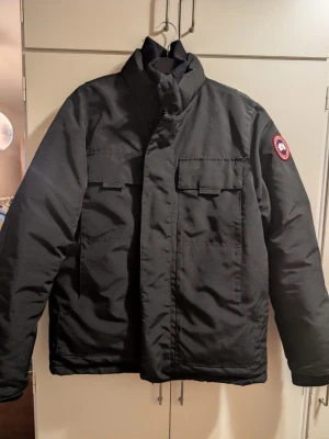 Svart dunjacka från Canada Goose - Svart dunjacka från Canada Goose med hög krage och två stora bröstfickor med lock. Klassisk patch på vänster ärm. Perfekt för kalla vinterdagar och riktigt varm. Snygg och stilren design som passar till det mesta. Varm vinterjacka, använd en säsong.