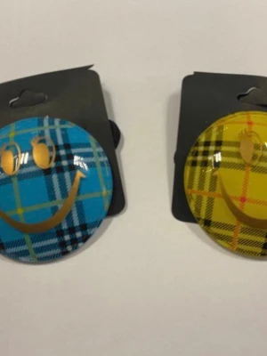 Hårsnoddar med rutigt smiley-motiv - Två elastiska hårsnoddar med stora runda dekorationer i blått och gult rutigt mönster. Båda har ett guldigt smileyface på framsidan. Perfekt för att ge din frisyr en färgglad och lekfull touch.