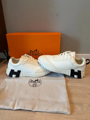 Vita  sneakers  - Säljer ett par vita  sneakers med chunky sula och svarta H-detaljer på sidorna. Skorna har en modern design med blandning av läder och textil, samt vita skosnören och svart hälflik. Perfekta för dig som gillar exklusiva och trendiga sneakers.